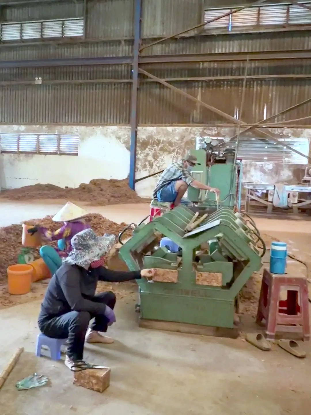 A worker in a sun hat sitting beside a green brick press, with coir bricks on the floor