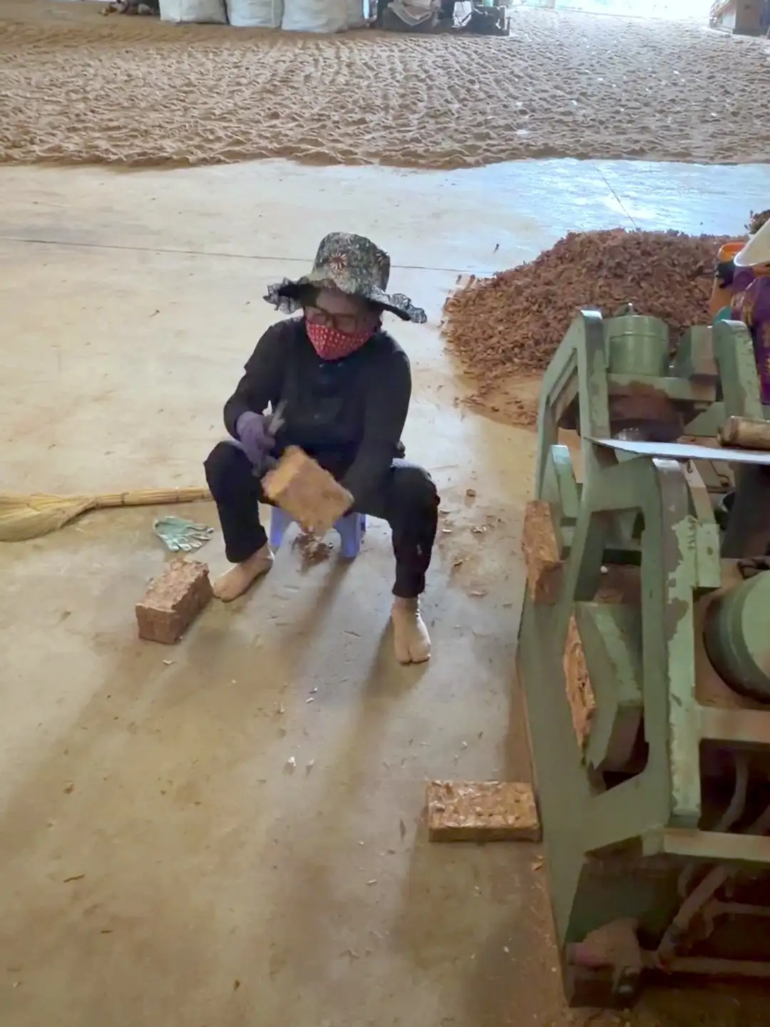 A worker in a sun hat and face mask seated beside a press, holding a freshly pressed coir brick